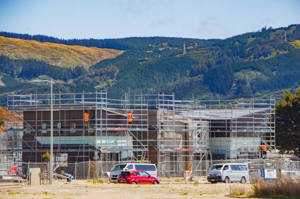 An under-contruction house surrounding by scaffolding, where construction workers stand in high-vis vests